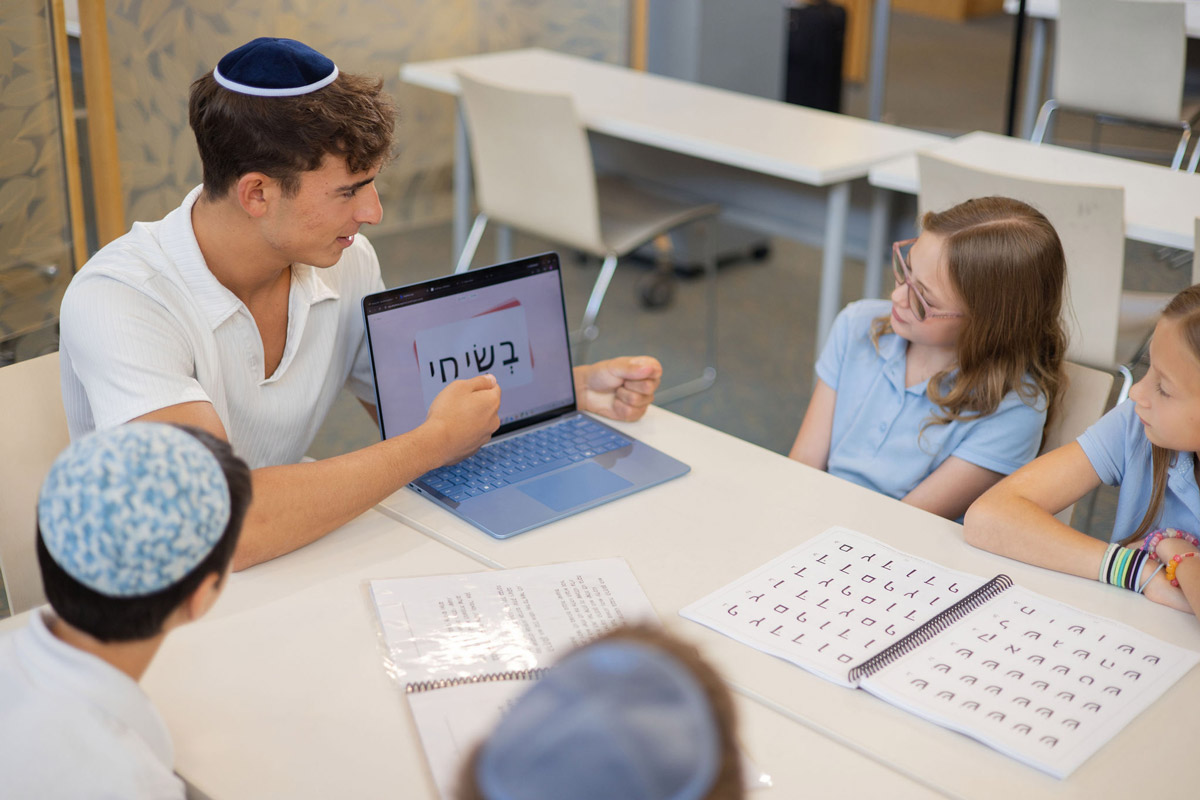 An adult teaches children gathered around a table with a laptop displaying Hebrew text and open workbooks.