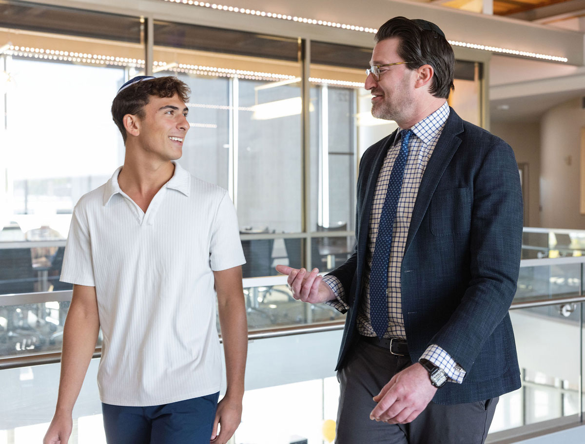 Two people walk through a modern building talking as they pass glass walls and office spaces.