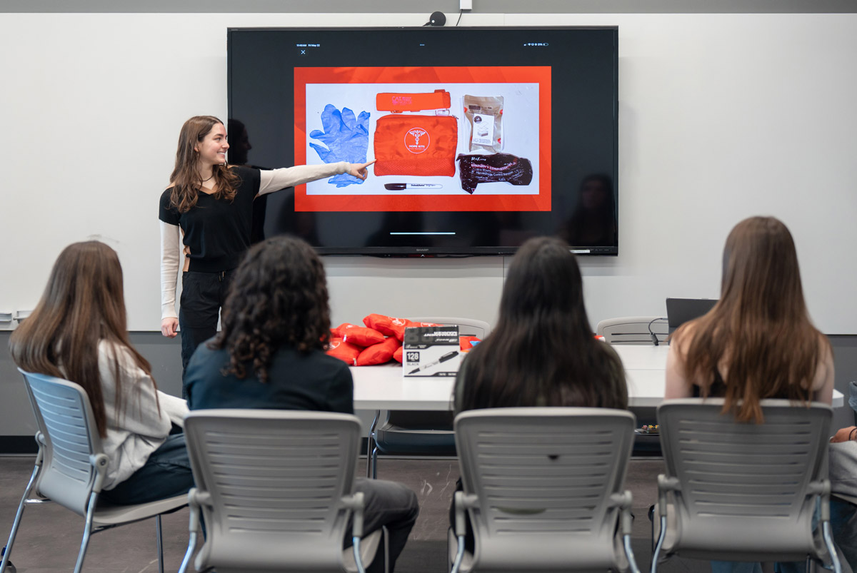 A person stands and points at a large screen while four others seated watch a presentation about items displayed.