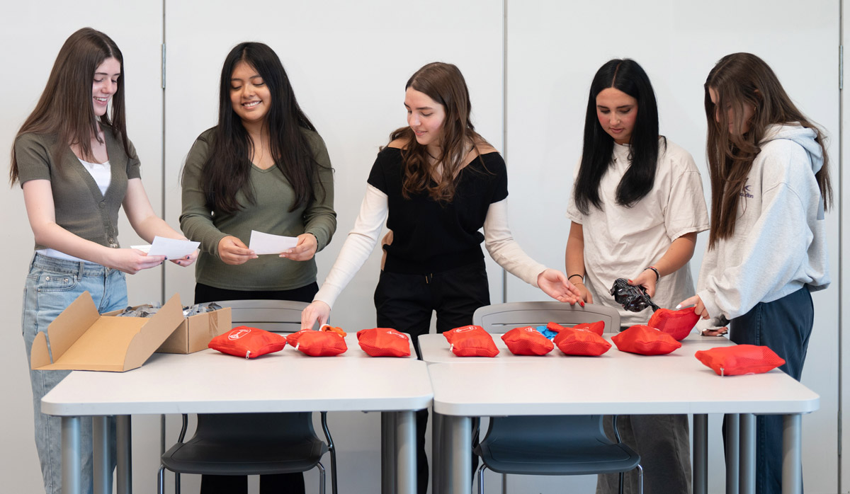 Five people stand around two tables sorting small red pouches and papers during a group activity.