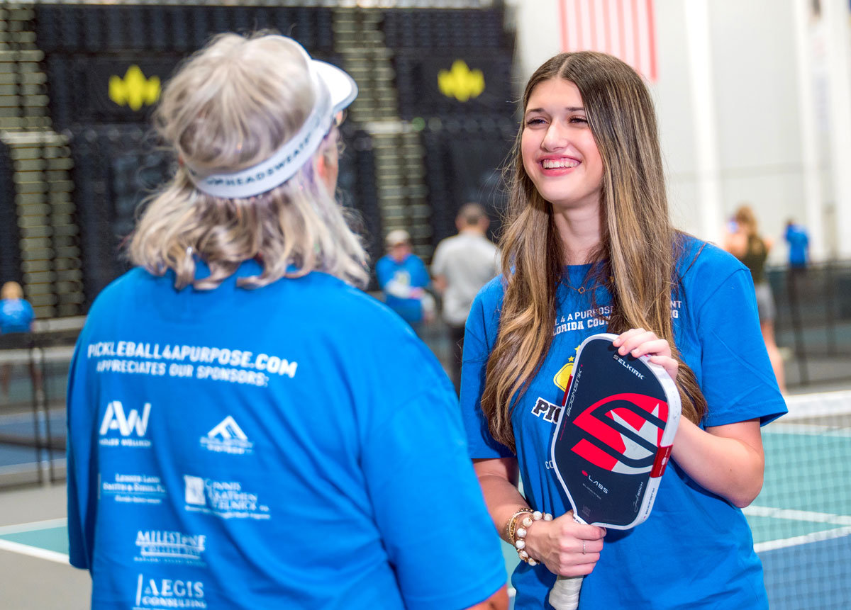 Teen holds a pickleball paddle while talking with another participant at an indoor pickleball event.