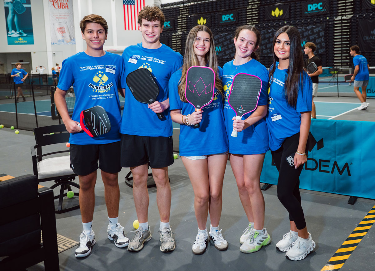 Five teens pose with pickleball paddles on an indoor court during a pickleball tournament.