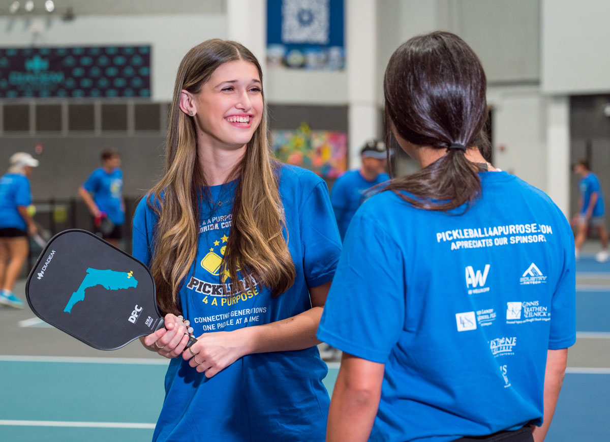 Teen holds a pickleball paddle while talking with another participant on an indoor pickleball court.