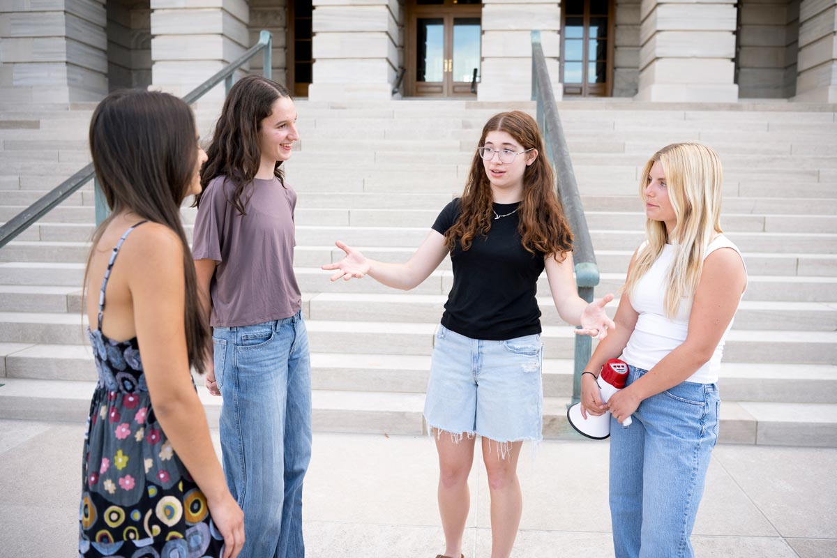 Four people stand talking on stone steps outside a large building as one gestures and another holds a megaphone.
