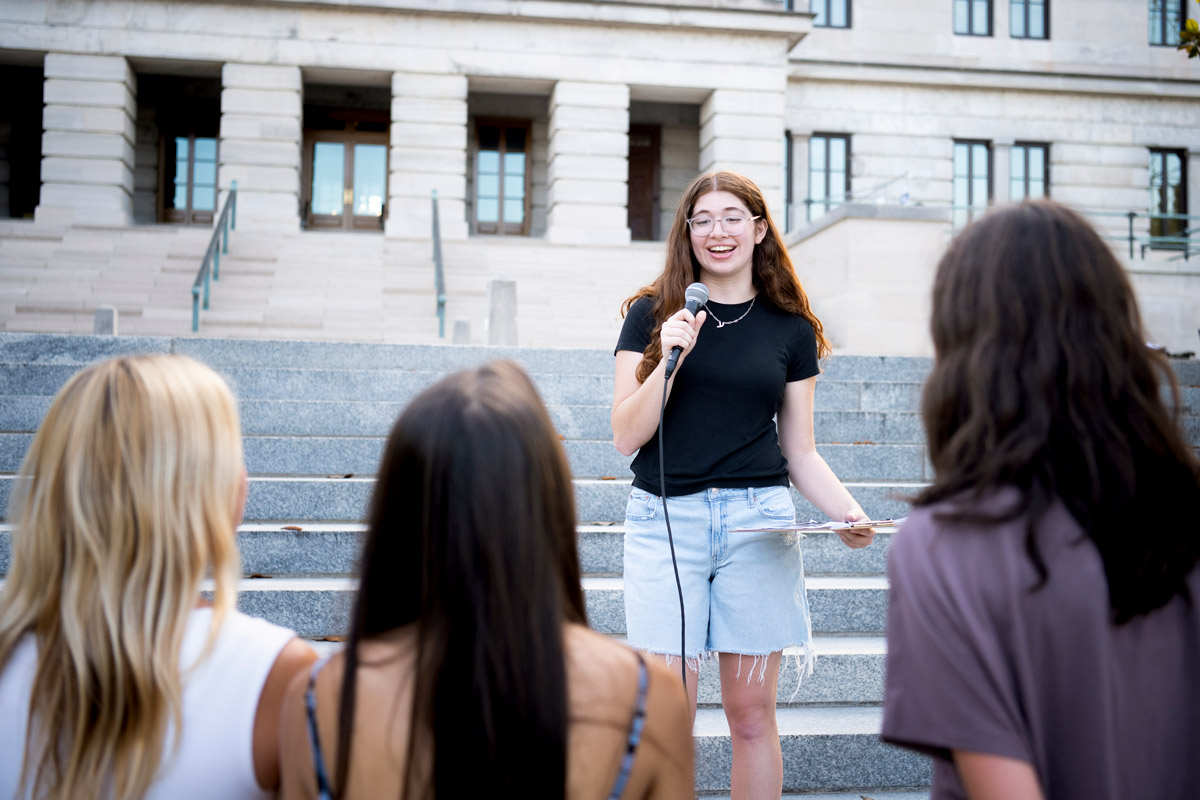 Person speaks on outdoor stone steps holding a microphone and clipboard as three people listen below.