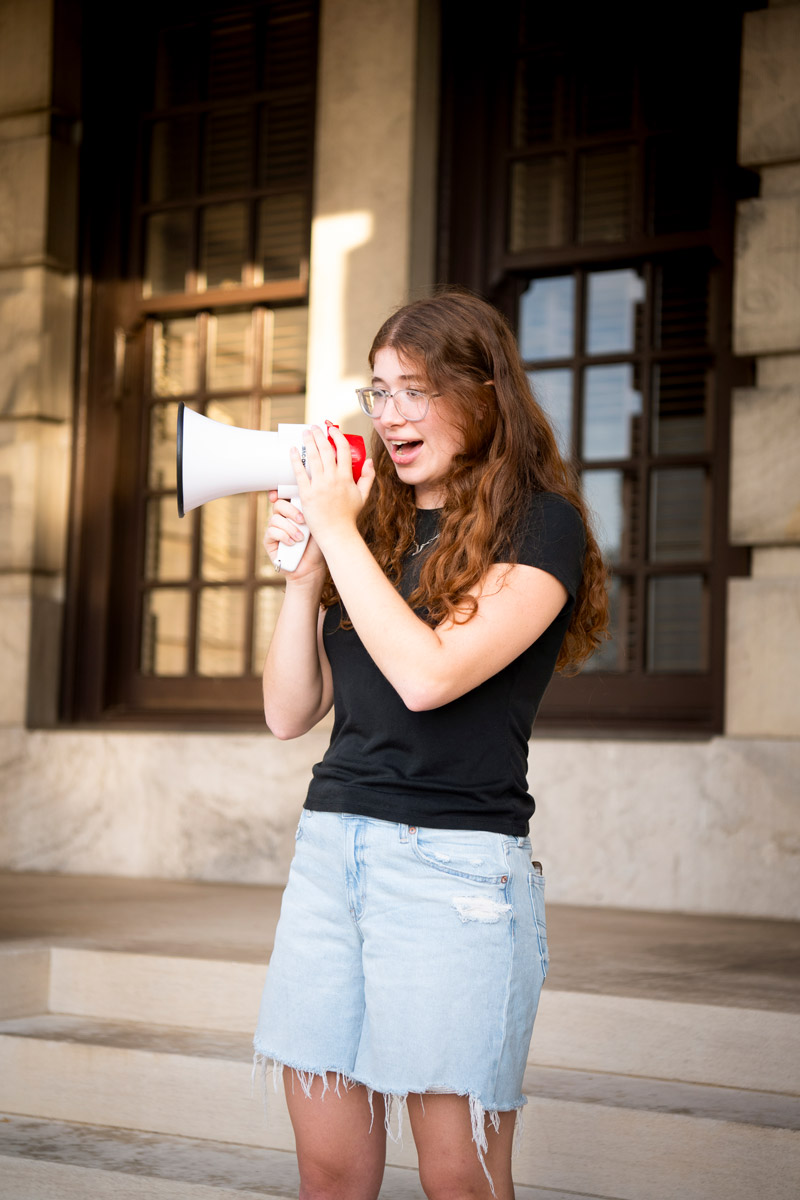 Person with long hair and glasses speaks into a megaphone while standing on stone steps outside a large building.