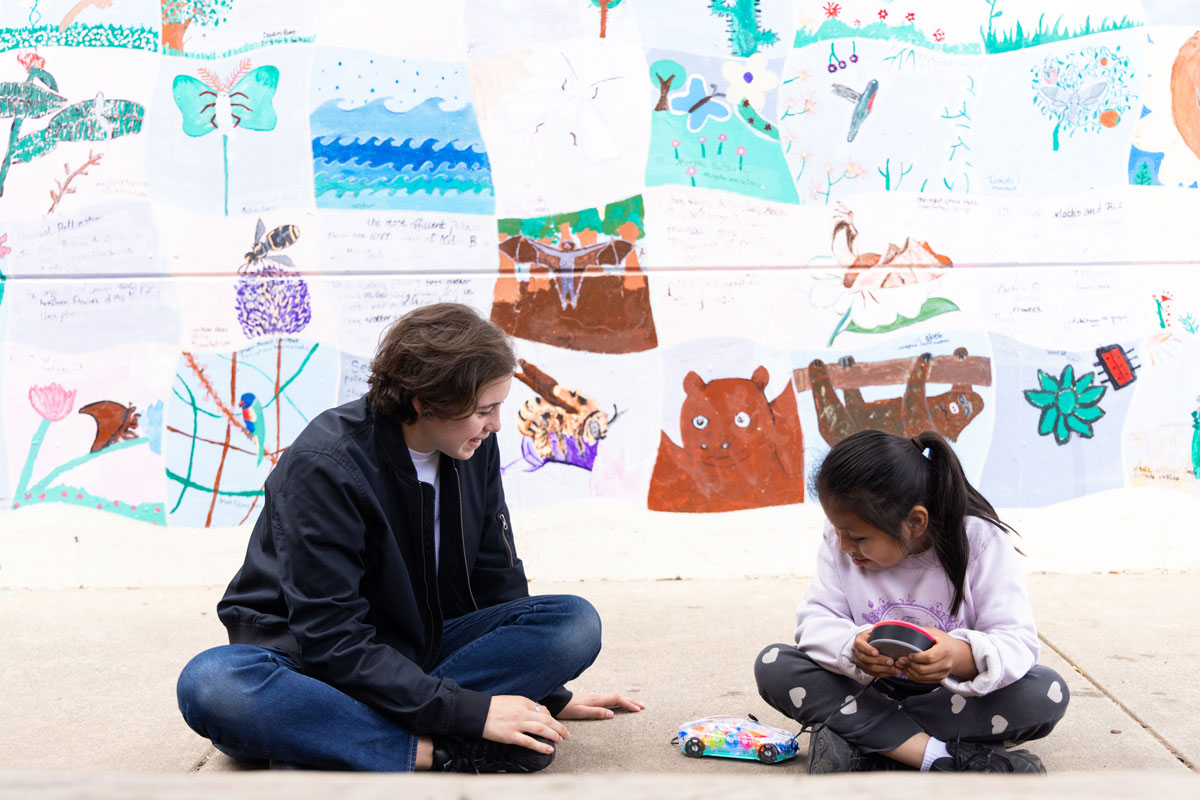 Two people sit cross‑legged in front of a colorful nature mural as one holds a round object and a toy car rests nearby.